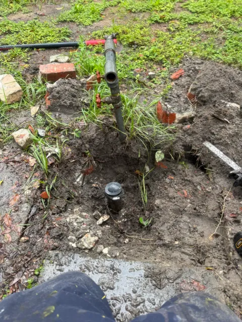 Dug-out hole in a yard with a capped pipe stub, next to a metal standpipe with red-handled valves