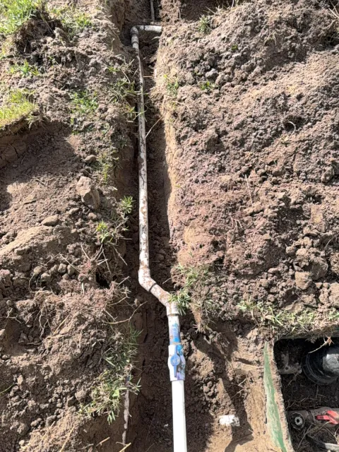 Open trench in a yard with a new white PVC water line and blue-handled valve next to a meter box